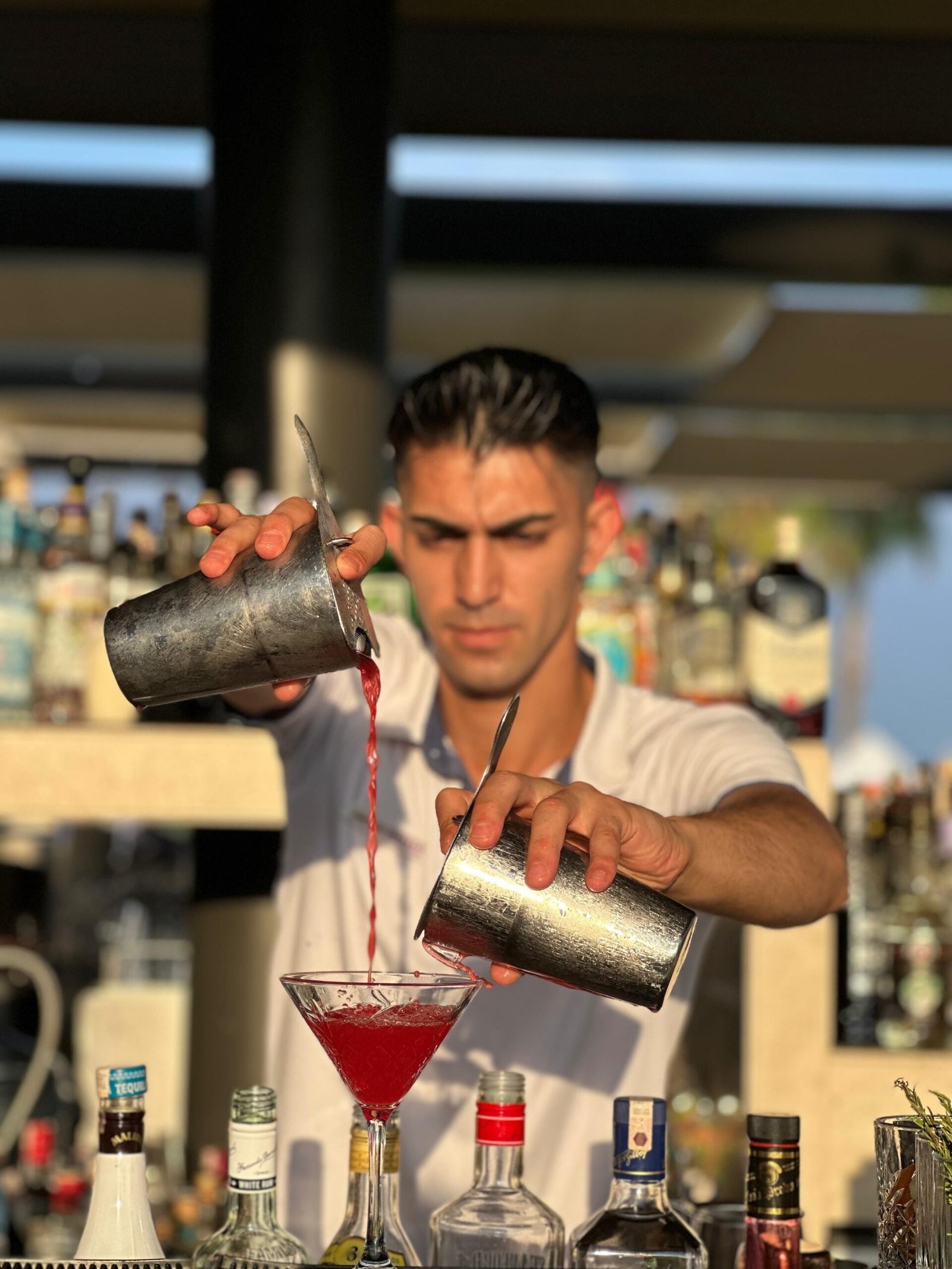 Home A bartender skillfully pours a red cocktail at an outdoor bar, showcasing mixology expertise.