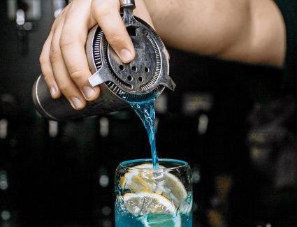pexels photo 33247511 33247511 A bartender pours a vibrant blue cocktail with lemon slices in a glass at a bar.